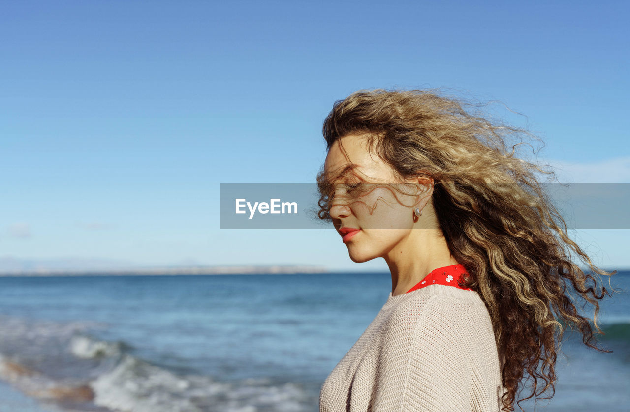 Side view of young woman with curly hair standing at beach against clear blue sky