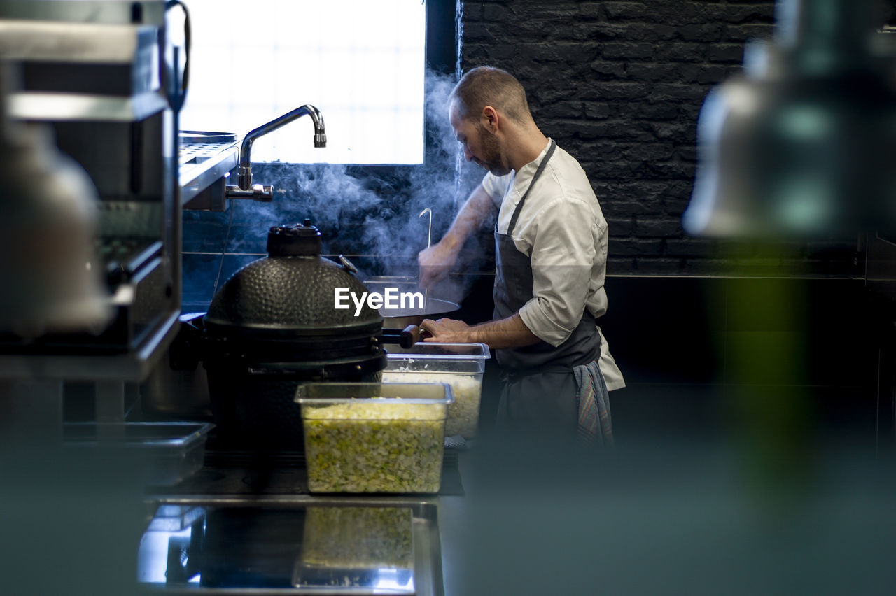 Young man in grey apron stirring big metal pan on restaurant kitchen