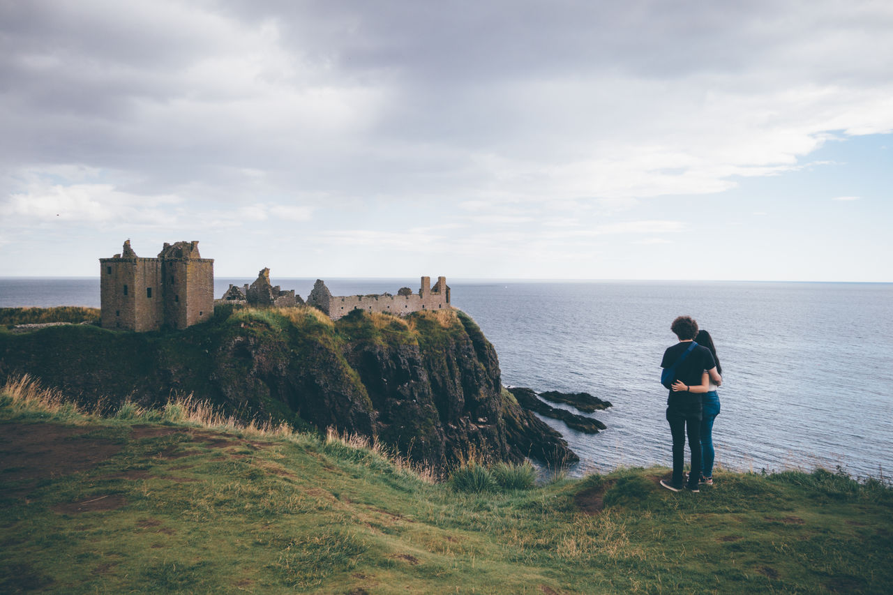 Rear view of couple standing by dunnottar castle against cloudy sky
