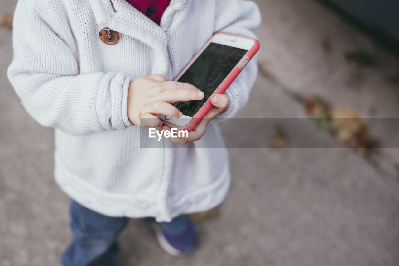 Low section of girl holding smart phone while standing on footpath