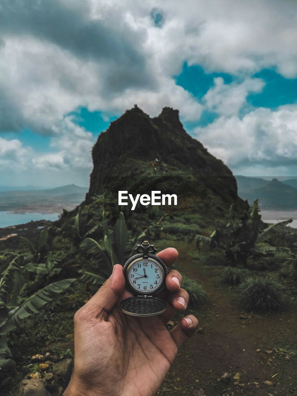 Cropped hand holding pocket watch near mountain against cloudy sky