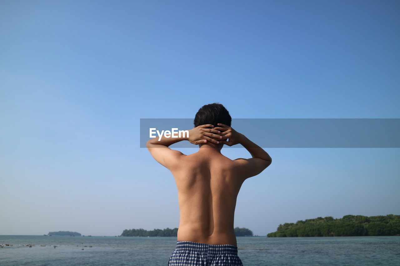 Rear view of shirtless man with hands behind head standing at beach against clear sky