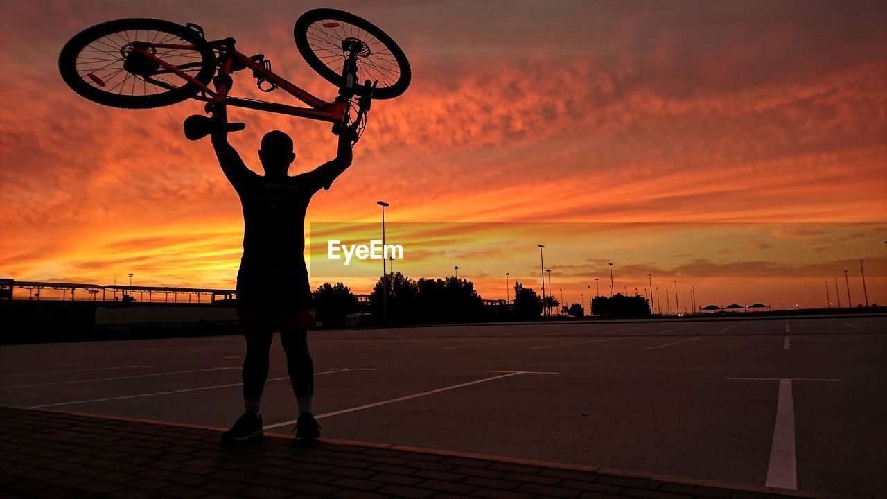 Silhouette man holding bicycle while standing at parking lot against cloudy sky during sunset