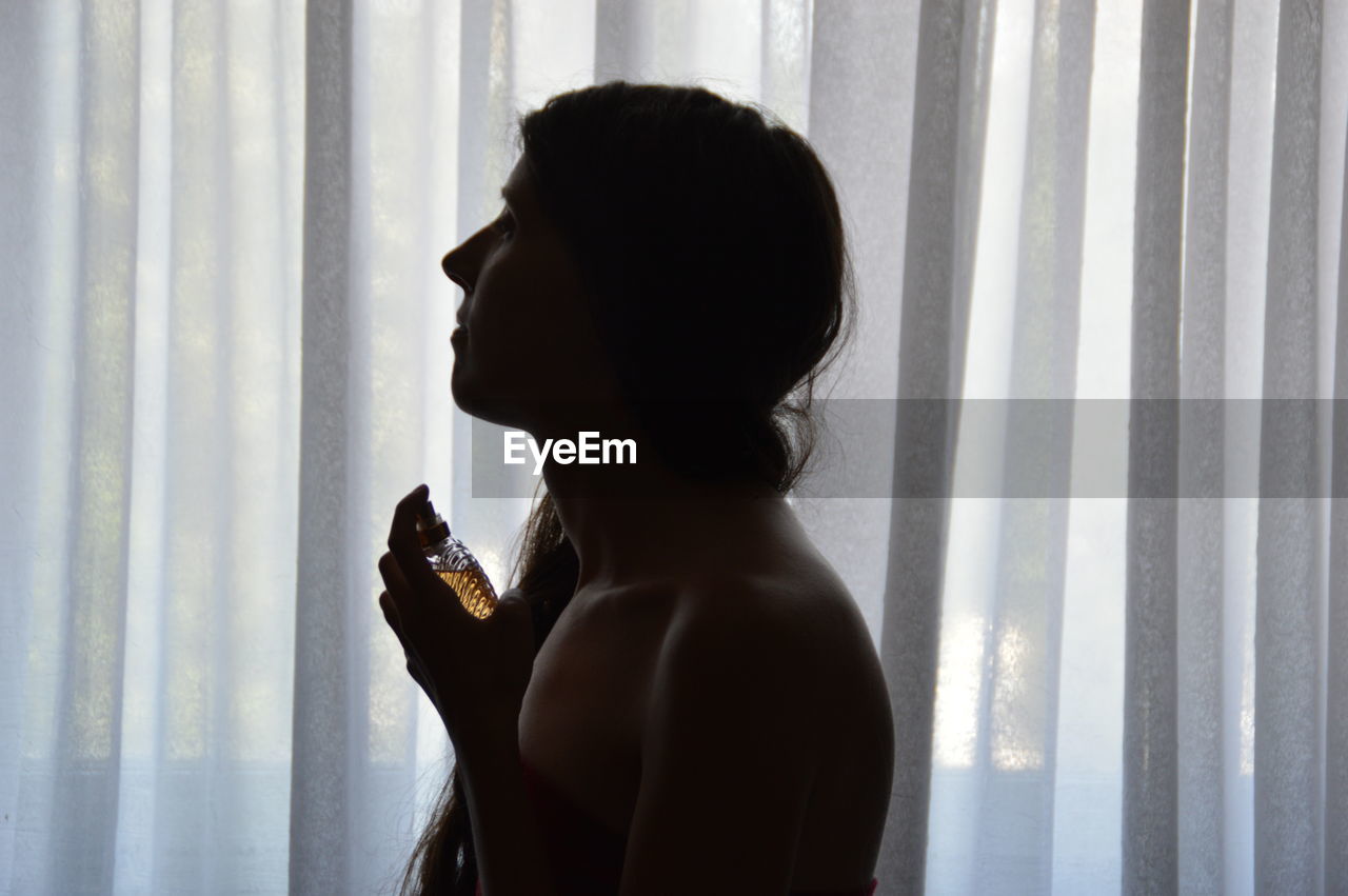 Side view of woman spraying perfume against curtain