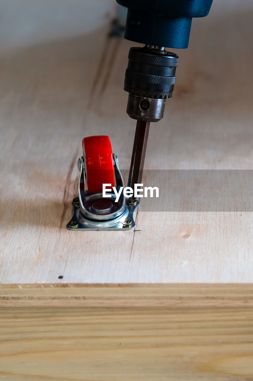 Close-up of electrical screwdriver fixing castor wheel on a wooden table.