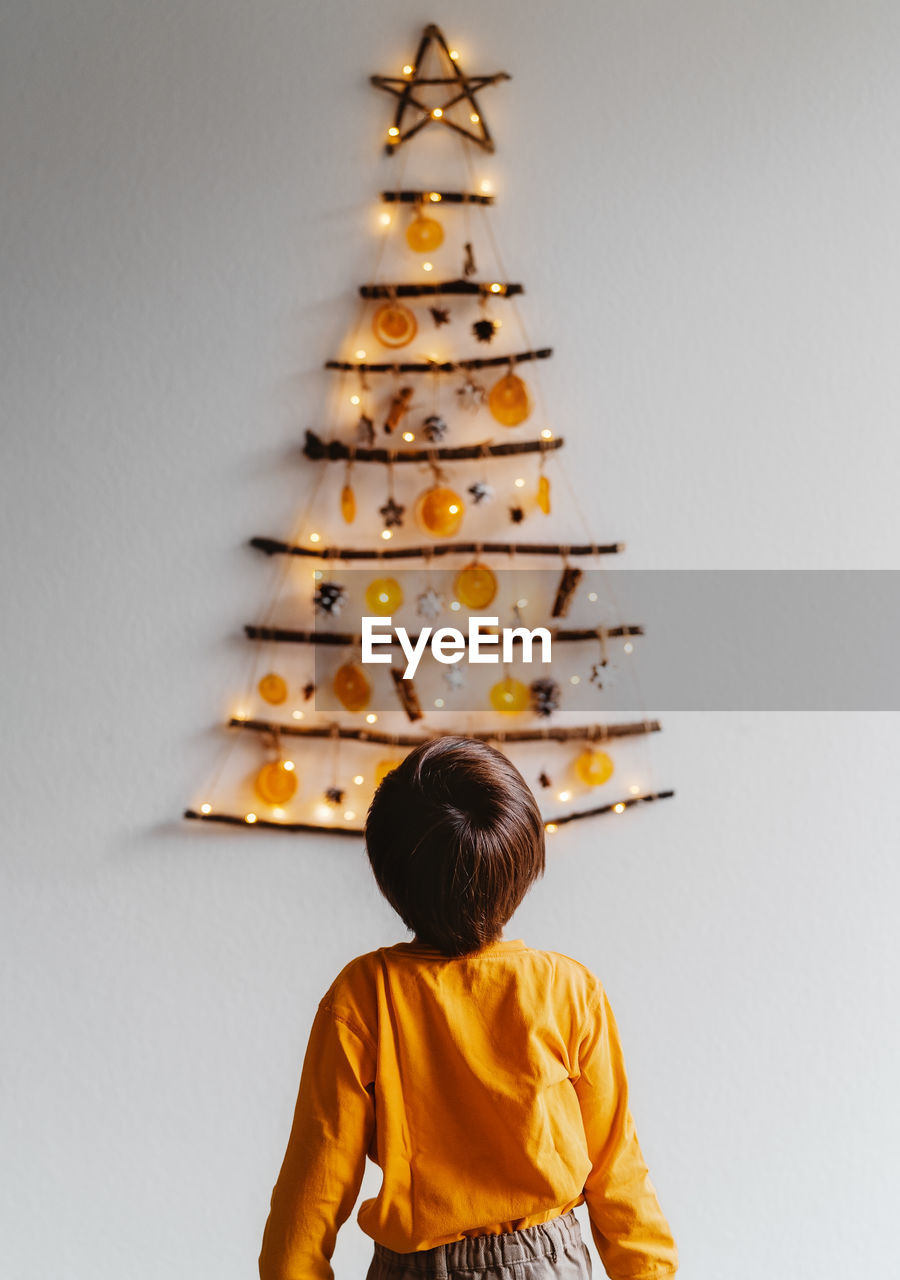 Rear view of child standing in front of handmade craft christmas tree hanging on wall. 