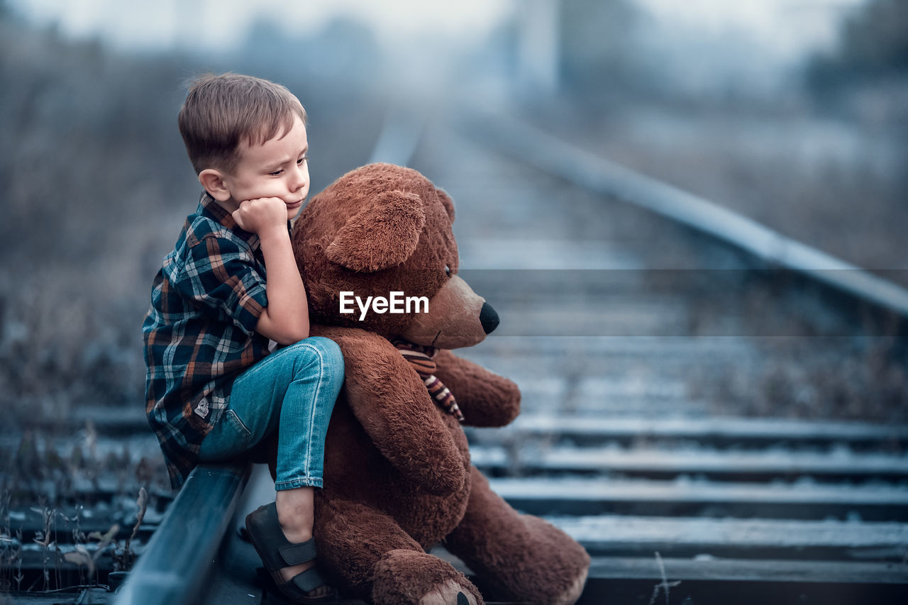 Side view of boy holding teddy bear while sitting on railroad track
