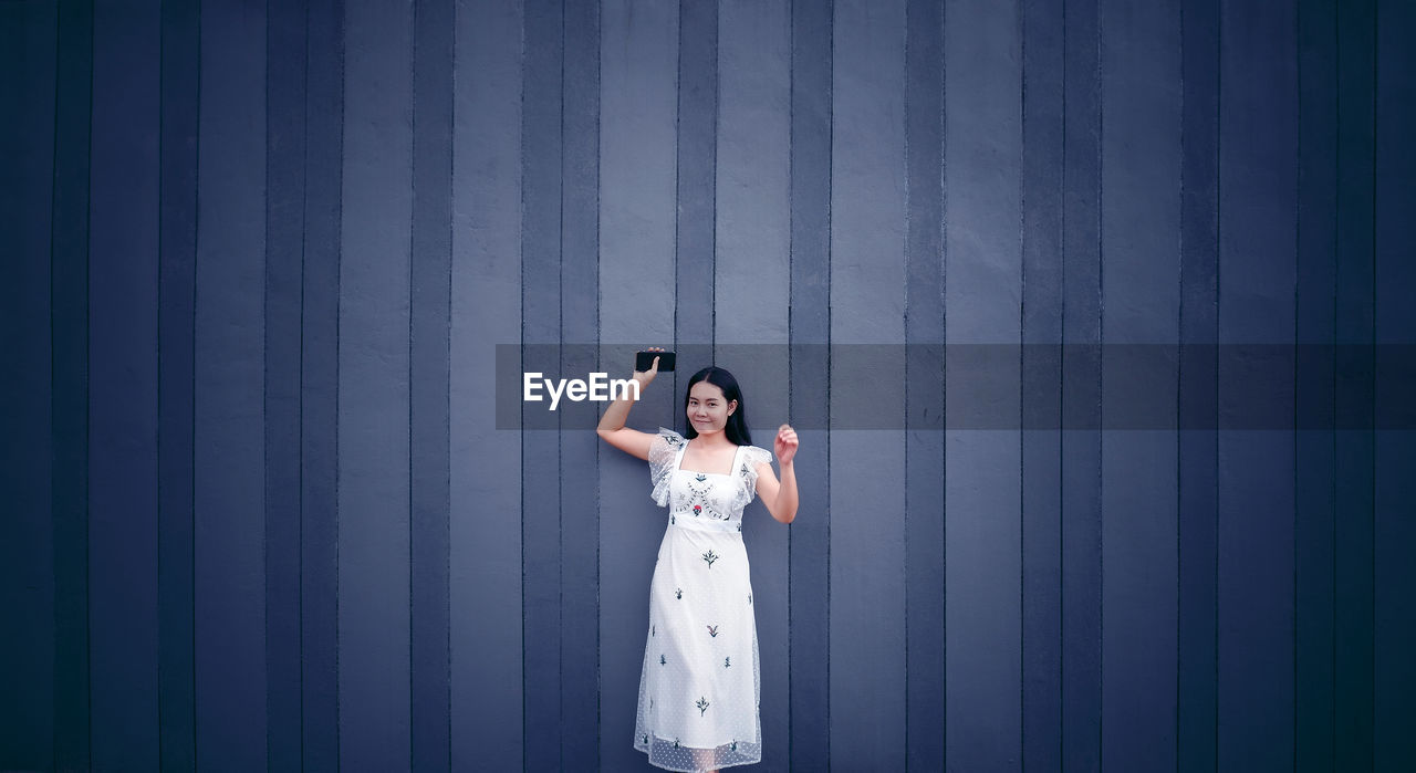 Portrait of woman holding mobile phone standing against wall