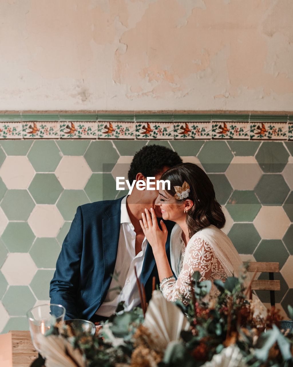 Anonymous ethnic man in suit whispering in ear of content beloved with closed eyes while sitting at restaurant table on wedding day