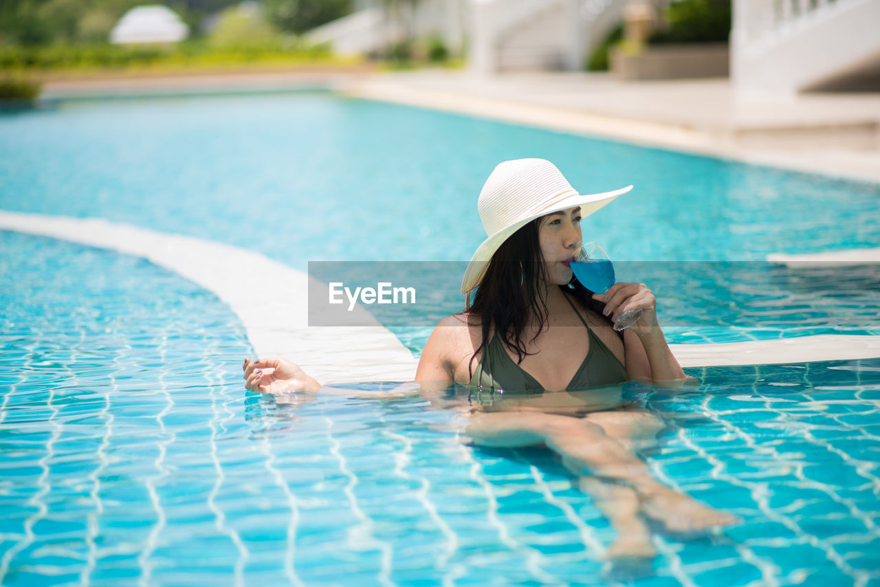 Young woman wearing hat drinking juice while relaxing in swimming pool 