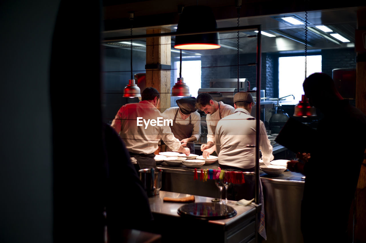 Young cooks men in aprons carefully serving meal in white ceramic dishes on restaurant kitchen