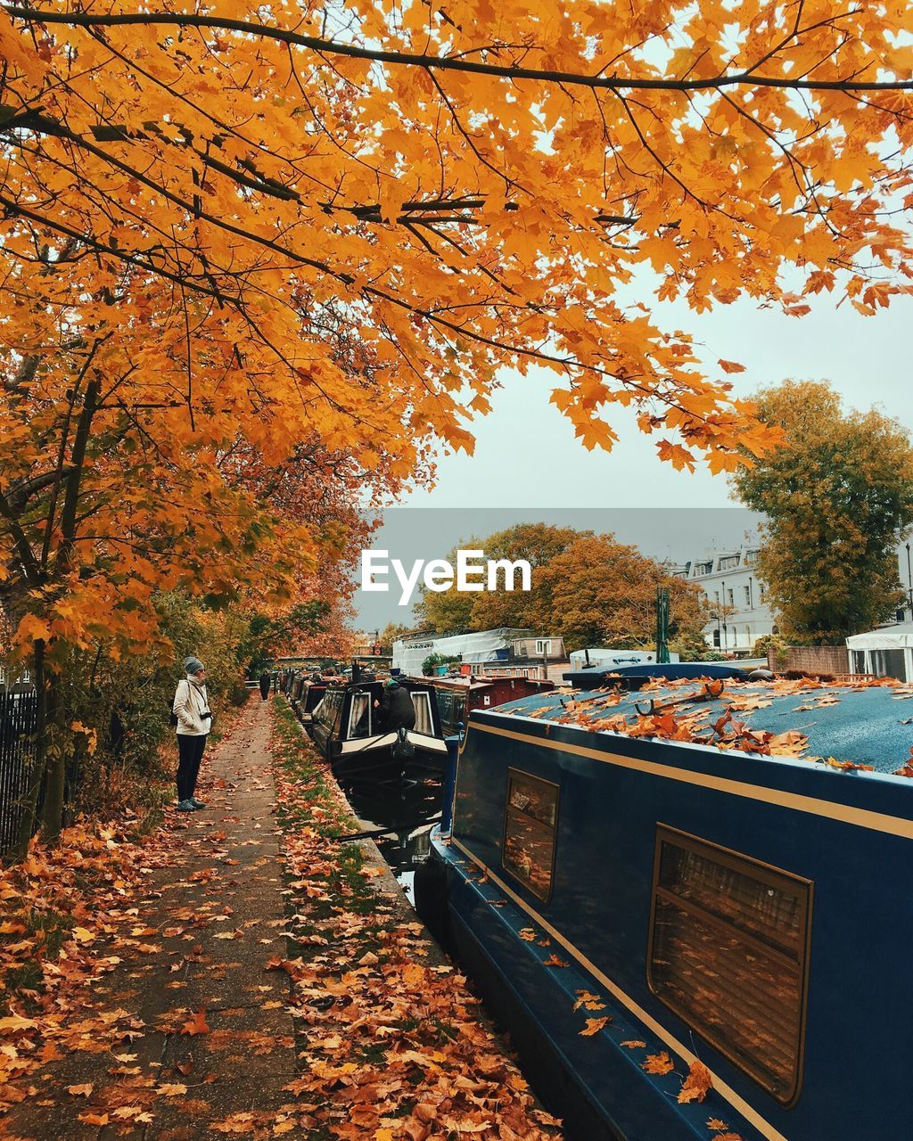 Woman standing on walkway by boats moored in canal during autumn