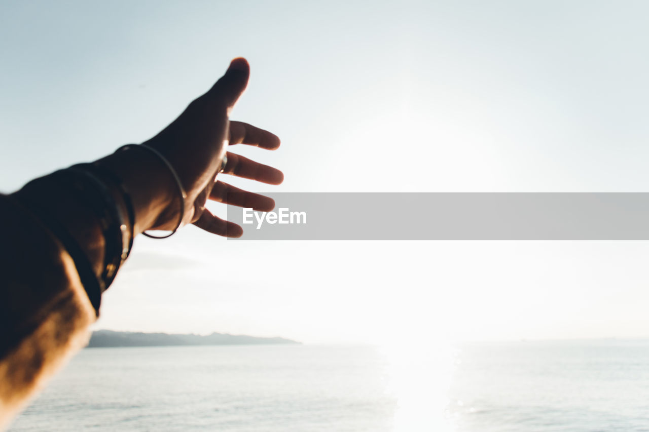 Cropped hand gesturing towards sea against sky during sunny day