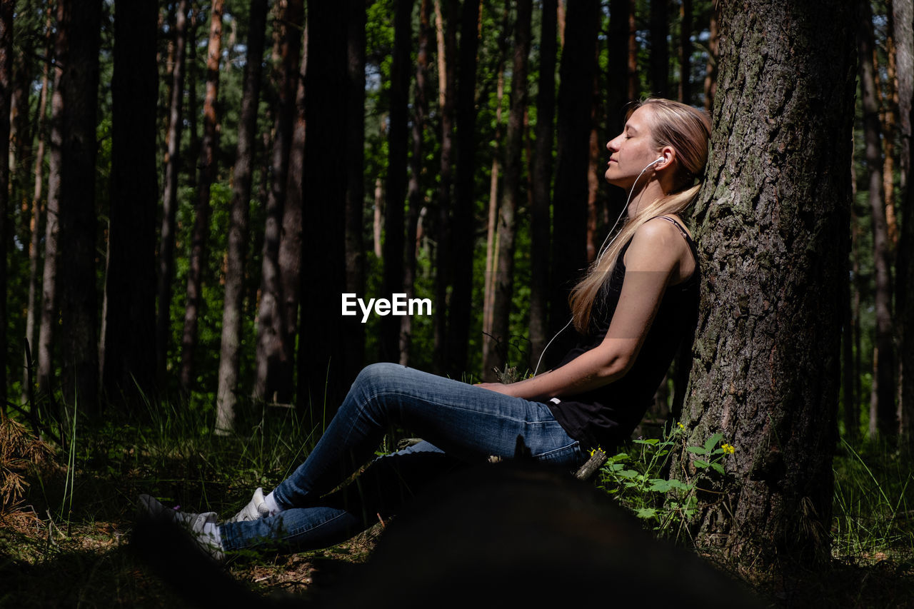 A girl in a forest sits leaning against a tree, headphones listening to music. positive emotion.