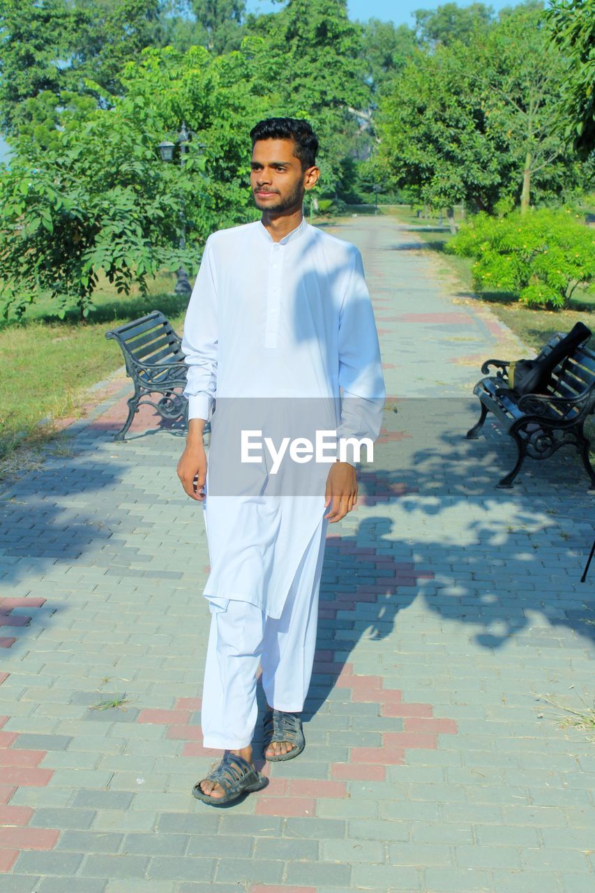 one person, full length, men, adult, plant, portrait, day, nature, young adult, clothing, tree, front view, looking at camera, casual clothing, lifestyles, standing, footpath, person, spring, leisure activity, sunlight, outdoors, footwear, smiling, walking, growth, shadow, beard, fashion, city