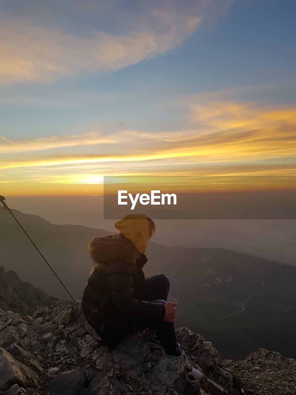 Side view of woman sitting on cliff against sky during sunset