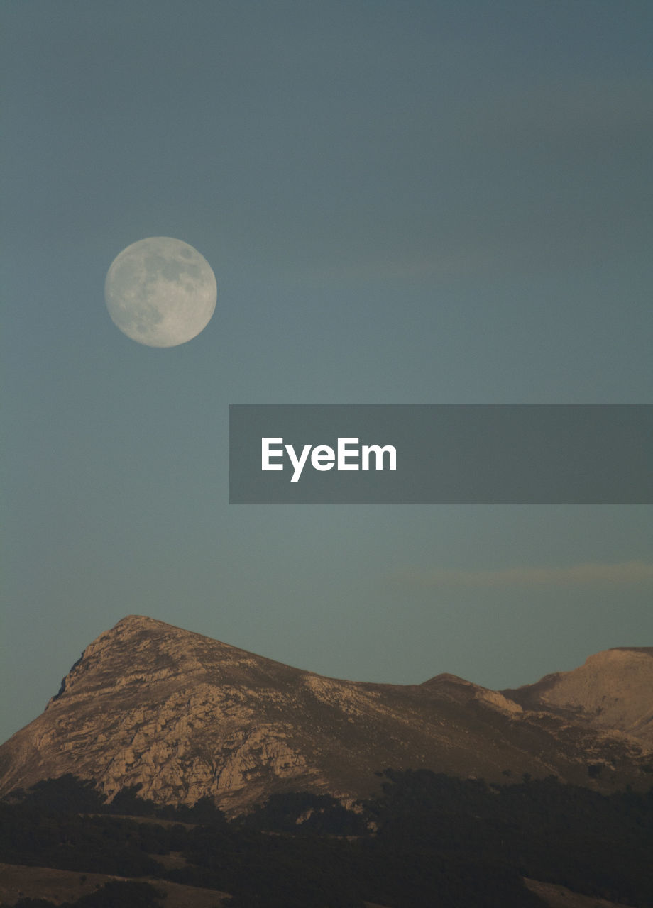 LOW ANGLE VIEW OF MOON IN MOUNTAINS AGAINST SKY