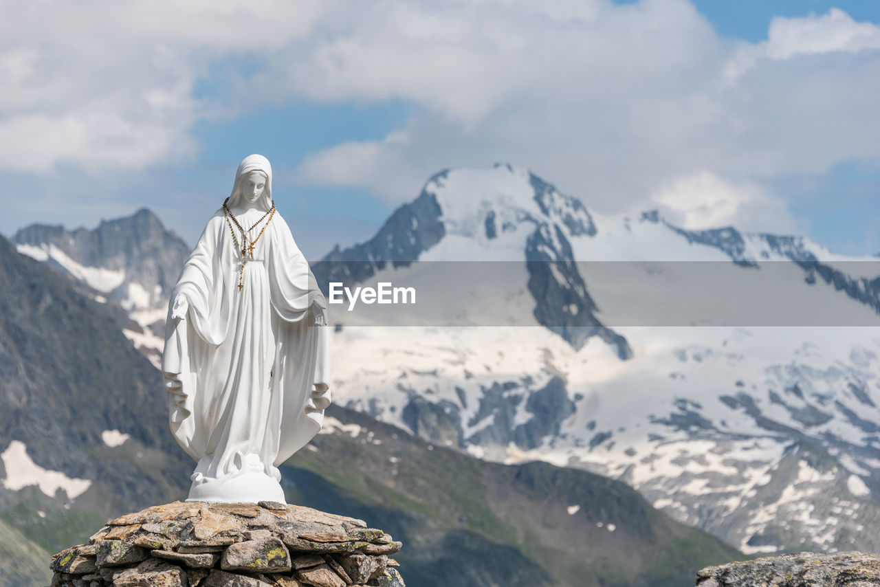 White statue of virgin mary, mother of god, placed on top of the mountain, blue sky, white clouds.