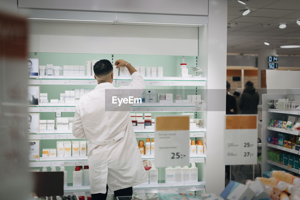 Rear view of male pharmacist arranging medicines on rack at store