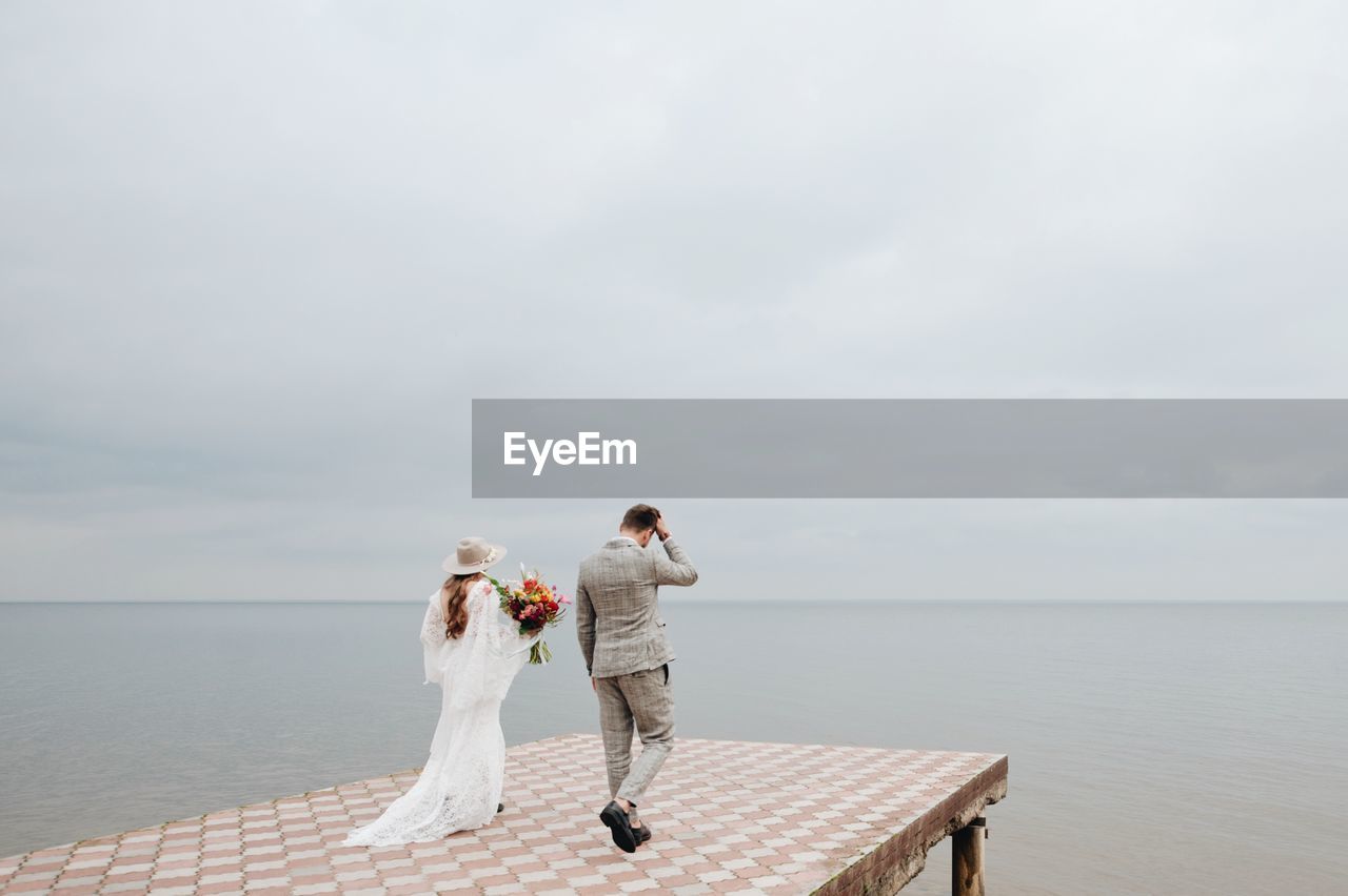 Bride and bridegroom walking on pier in sea during wedding ceremony