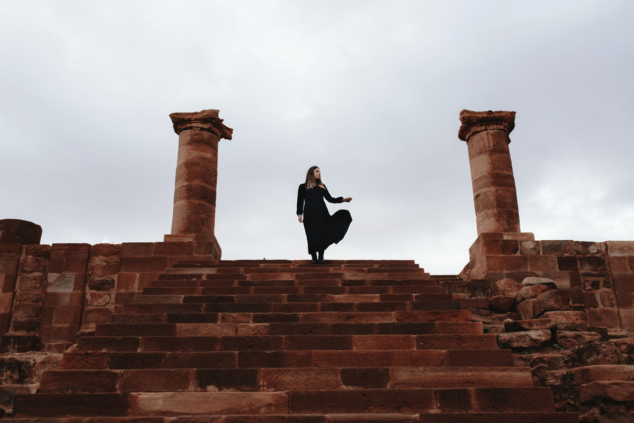 Low angle of unrecognizable young female traveler in maxi black dress standing on old stairs near destructed stone columns of great temple against cloudy sky in petra