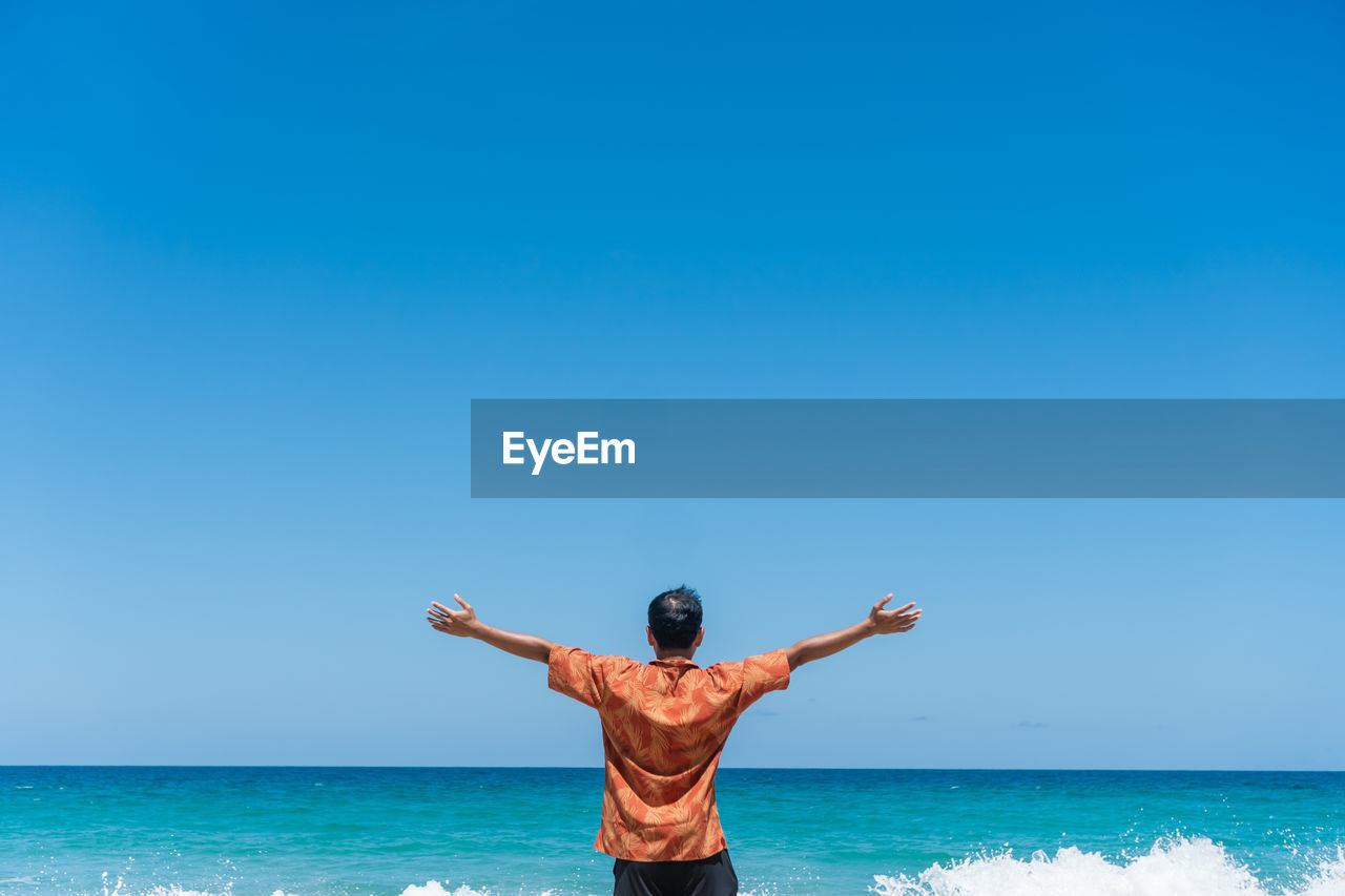 Rear view of man with arms outstretched standing at beach against sky