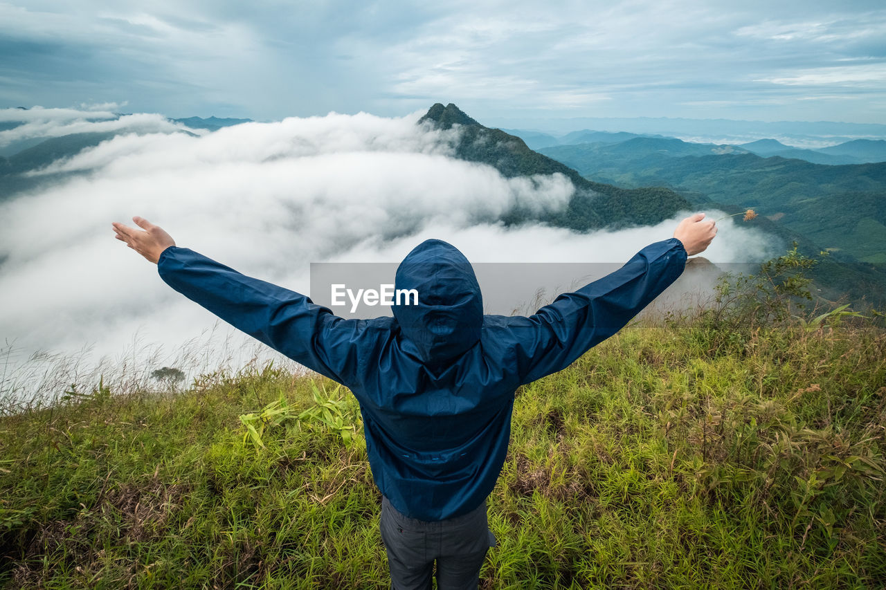 Rear view of person with arms outstretched looking at landscape against cloudy sky
