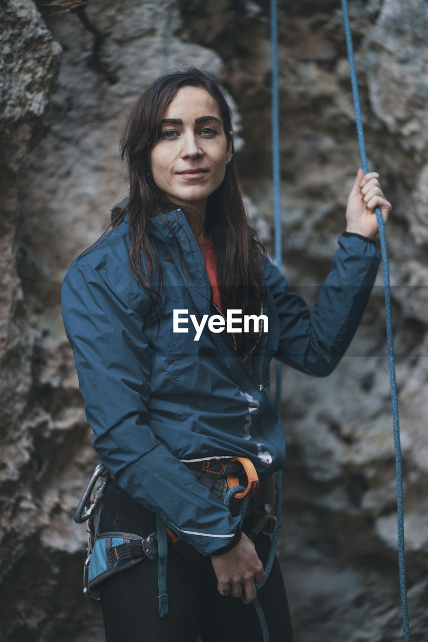 Portrait of young woman holding climbing rope while standing by mountain