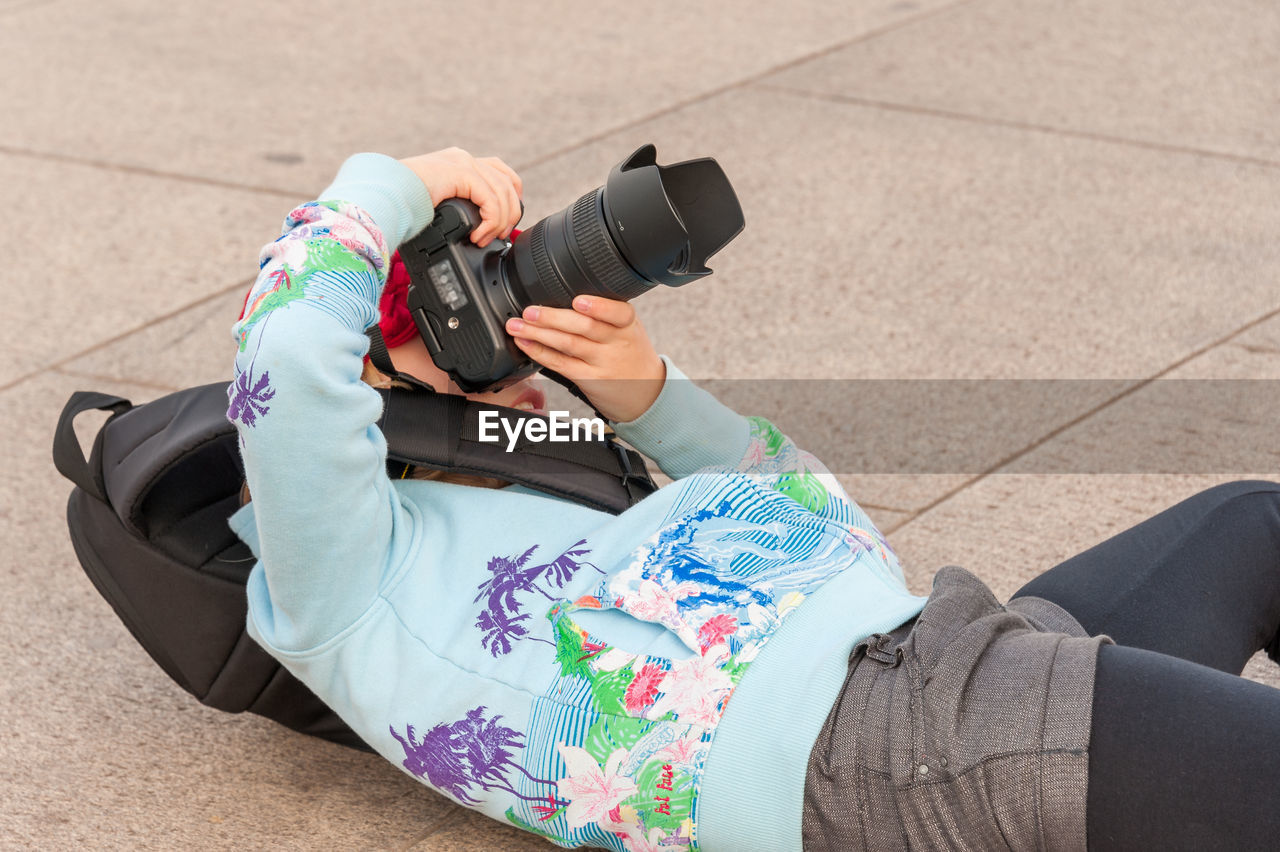 Very young female photographer lays on her back while using large dslr camera to get perfect angle.