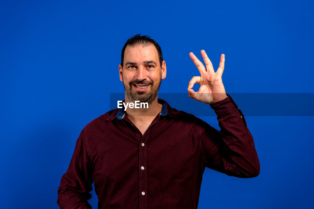 blue, one person, portrait, men, adult, sign language, studio shot, looking at camera, smiling, emotion, gesturing, happiness, waist up, colored background, blue background, person, front view, hand, cheerful, positive emotion, casual clothing, standing, copy space, indoors, hand sign, finger, sky, beard