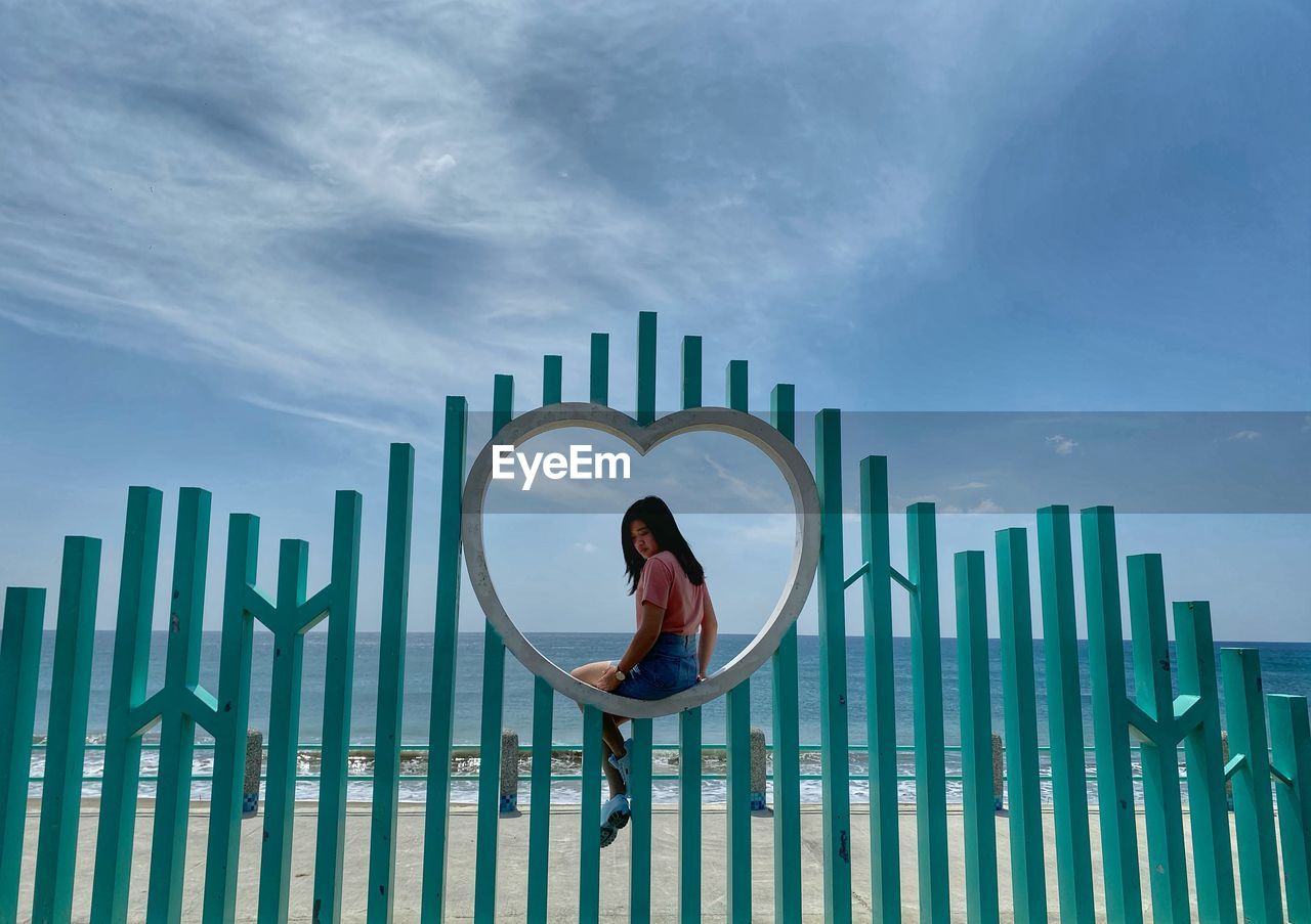 Woman sitting on heart shaped fence at beach against sky