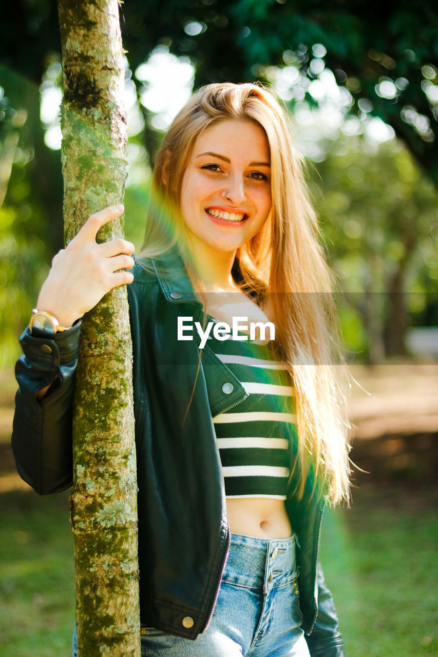 Portrait of smiling young woman standing by tree trunk in park