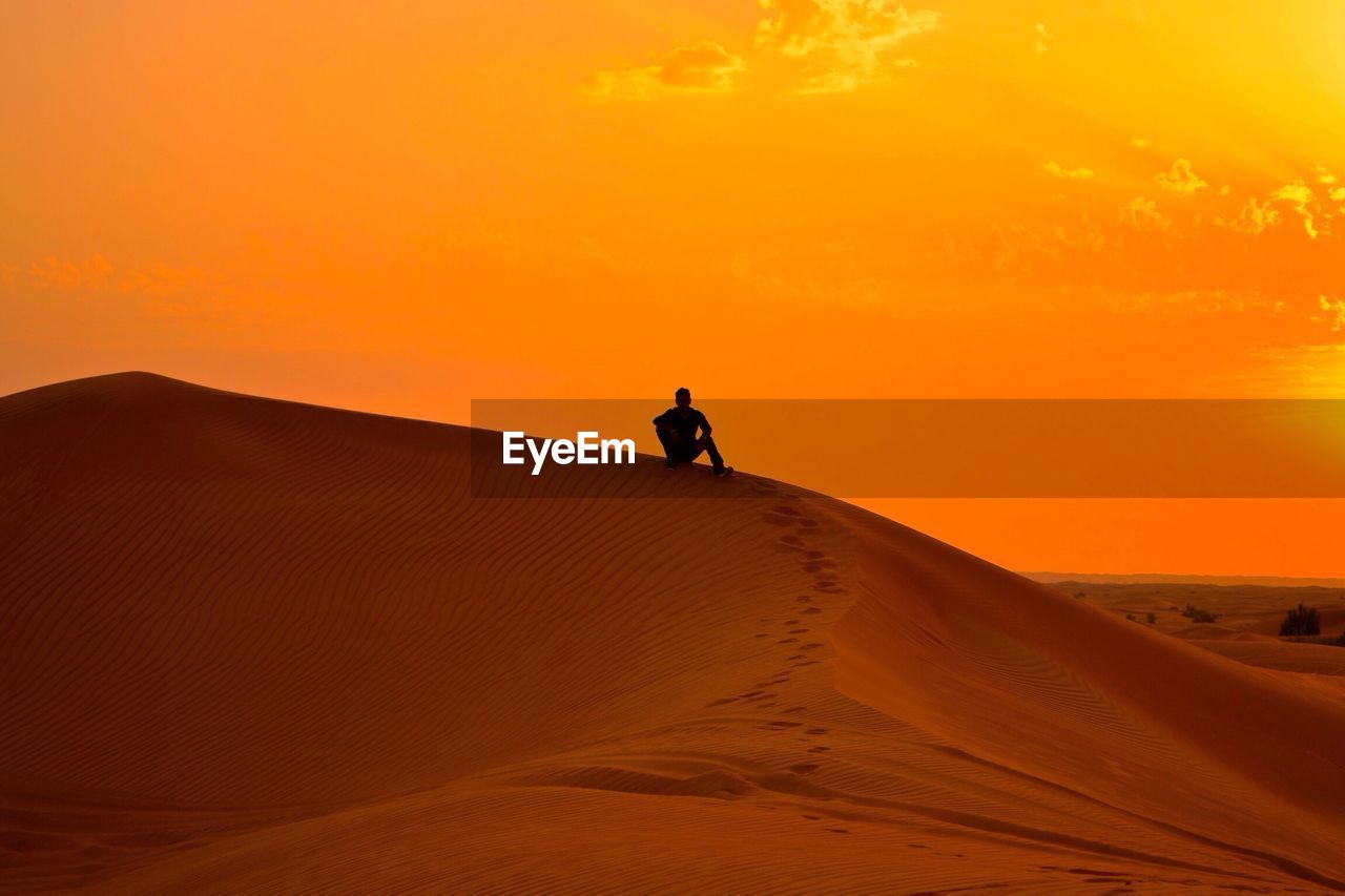 Silhouette man sitting sand dune in desert against orange sky