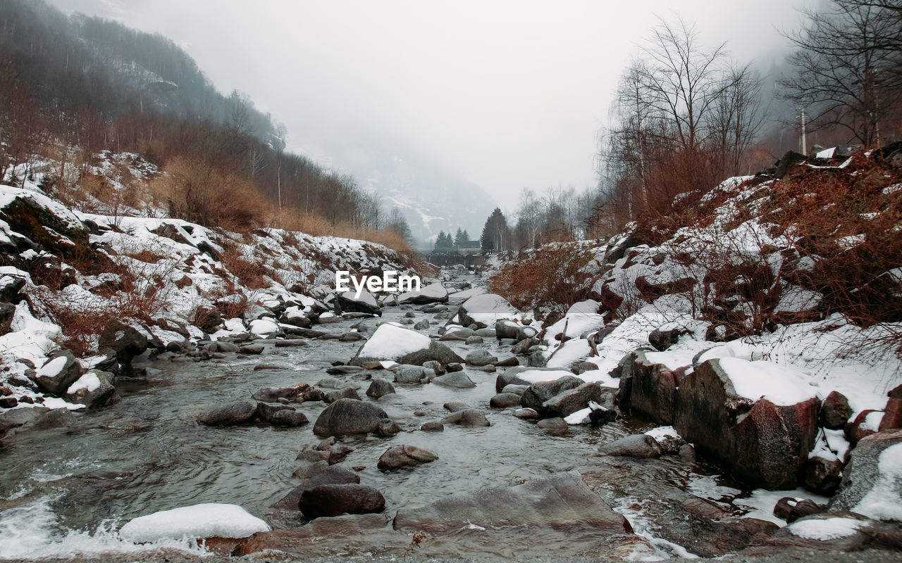 Scenic view of river flowing amidst rocks and bare trees in forest during winter