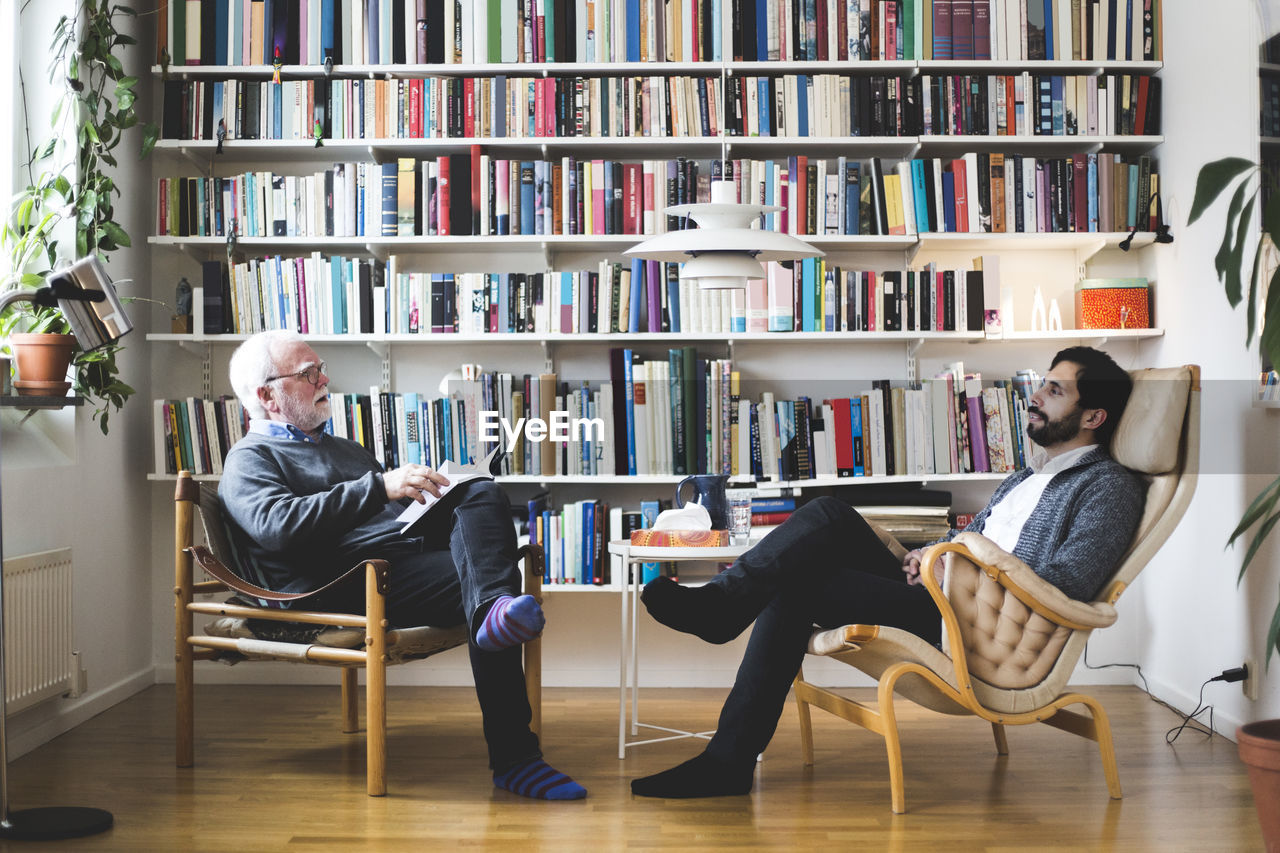 Therapist communicating with man while sitting by book shelf at home office