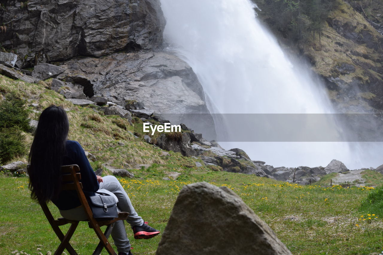 Rear view of woman sitting on chair while looking at waterfall