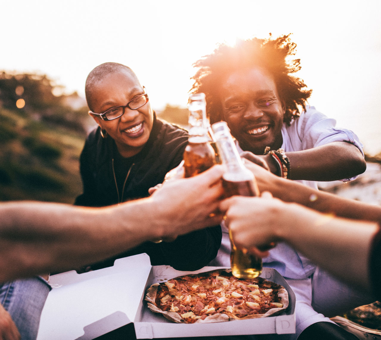 Friends toasting beer bottles while sitting against clear sky during sunset