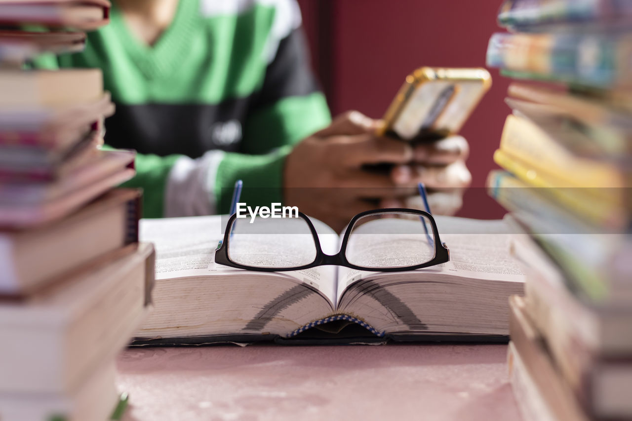 Selective focus on book and glasses through the stack of books while human hands with mobile phone 
