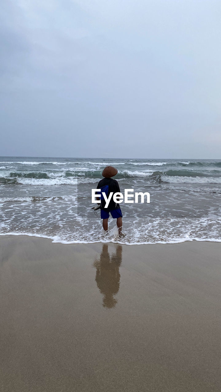 REAR VIEW OF BOY ON BEACH AGAINST SKY