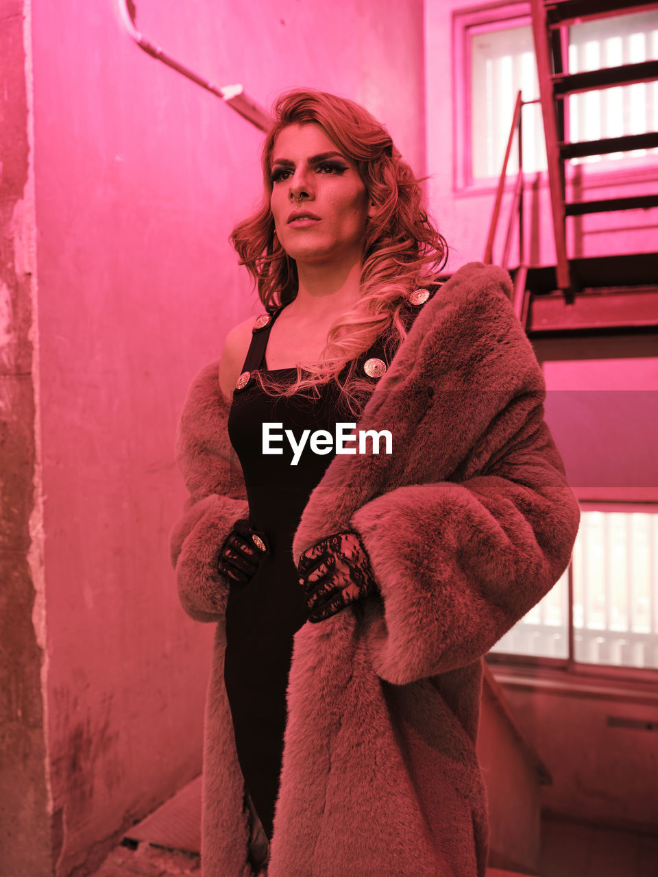 Stylish drag queen in elegant dress and fur coat looking away while standing near staircase under red light inside grungy building