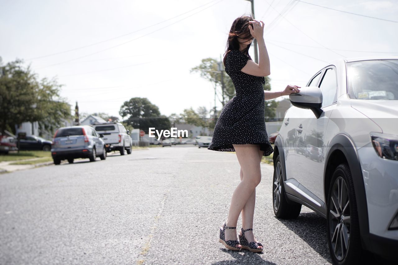 Side view of mid adult woman standing by car on road against sky during sunny day