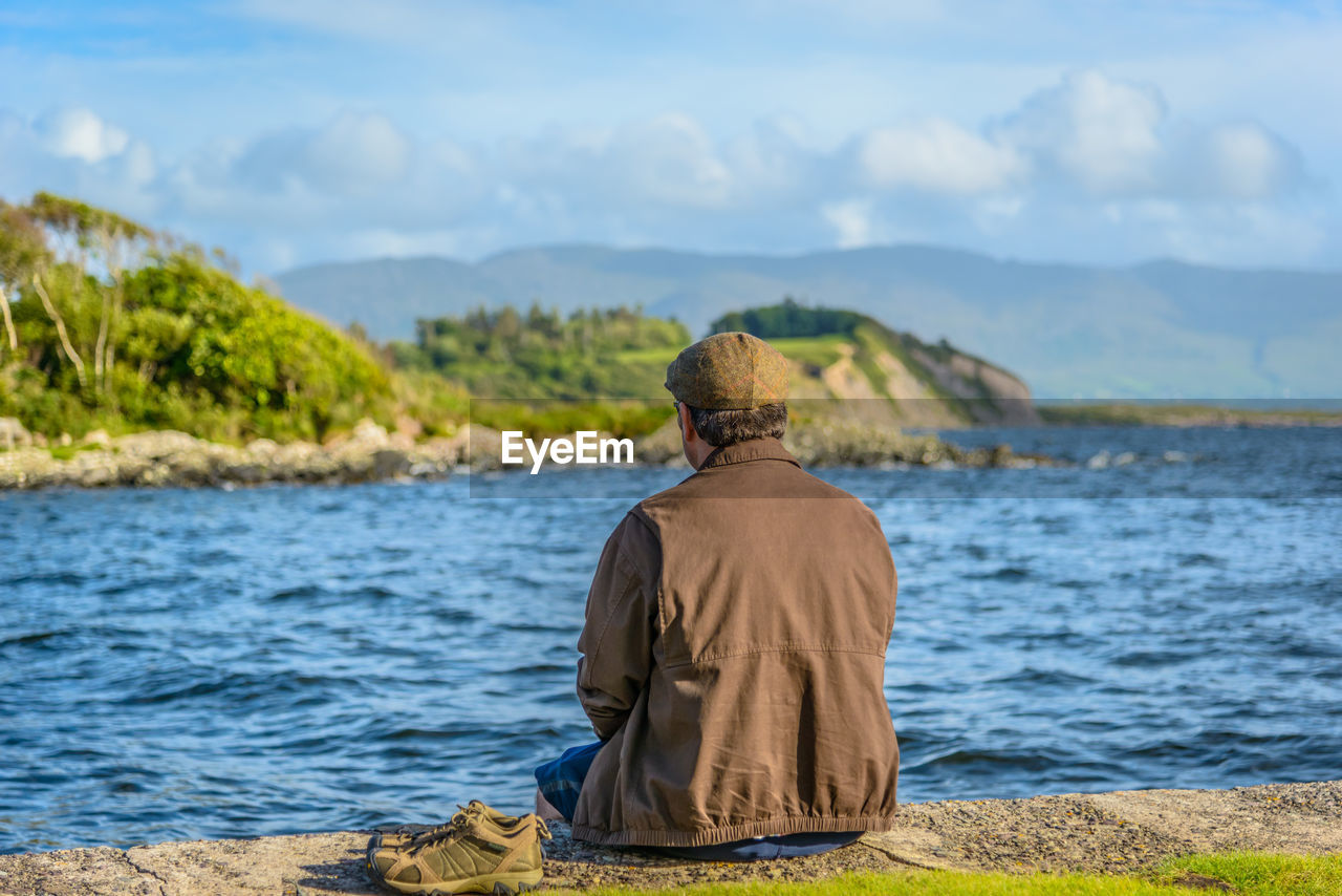 Rear view of man sitting at lakeshore against sky