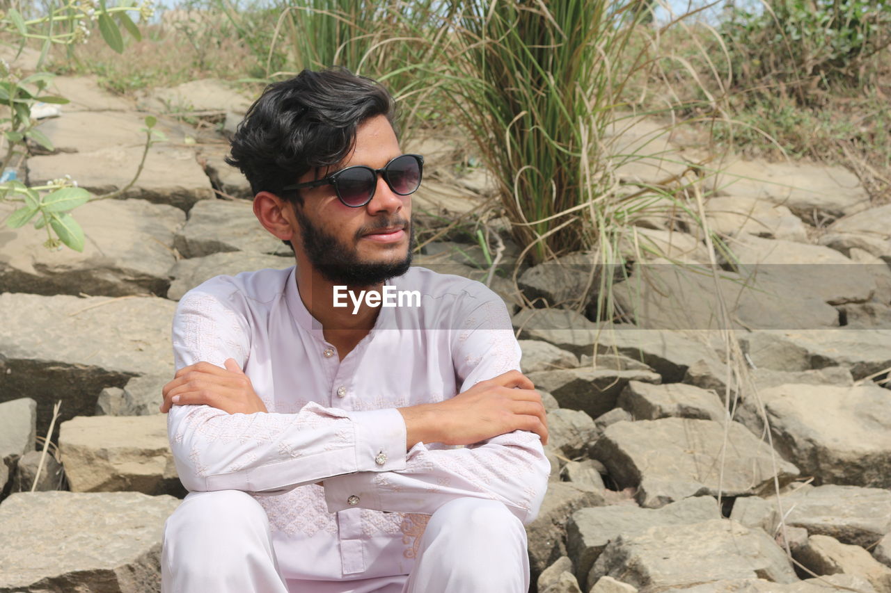 sunglasses, one person, sitting, young adult, glasses, beard, fashion, leisure activity, adult, men, casual clothing, facial hair, nature, front view, rock, lifestyles, relaxation, day, person, three quarter length, land, sunlight, outdoors, looking, clothing, looking away, portrait, vacation, trip, plant, black hair