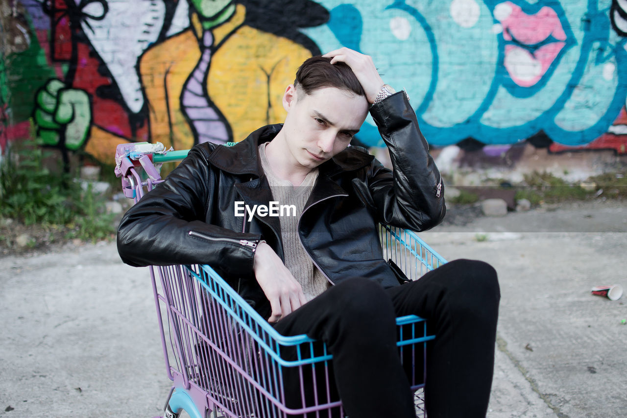 Young man sitting in shopping cart against graffiti on wall