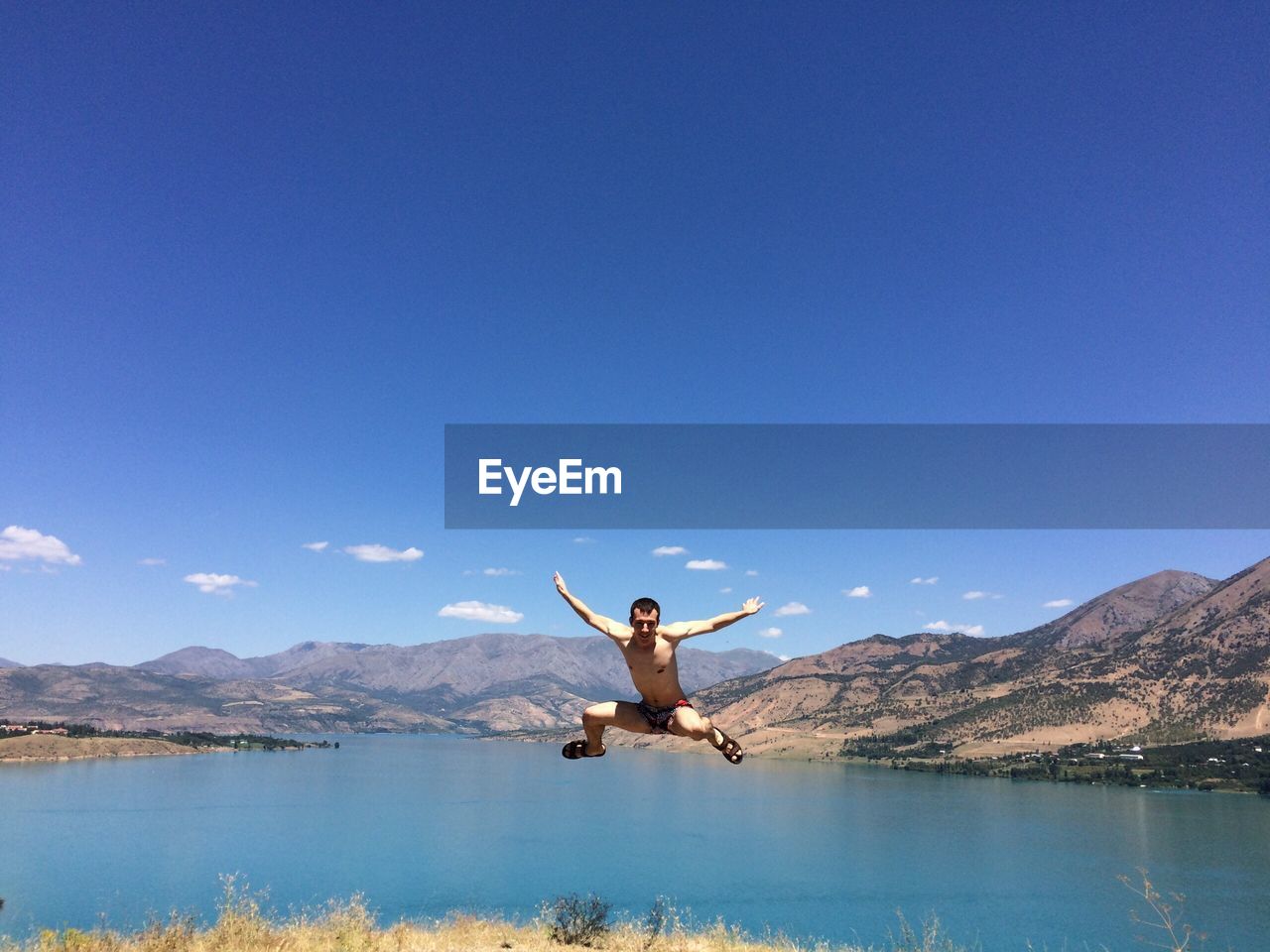 Shirtless man jumping at lake against sky