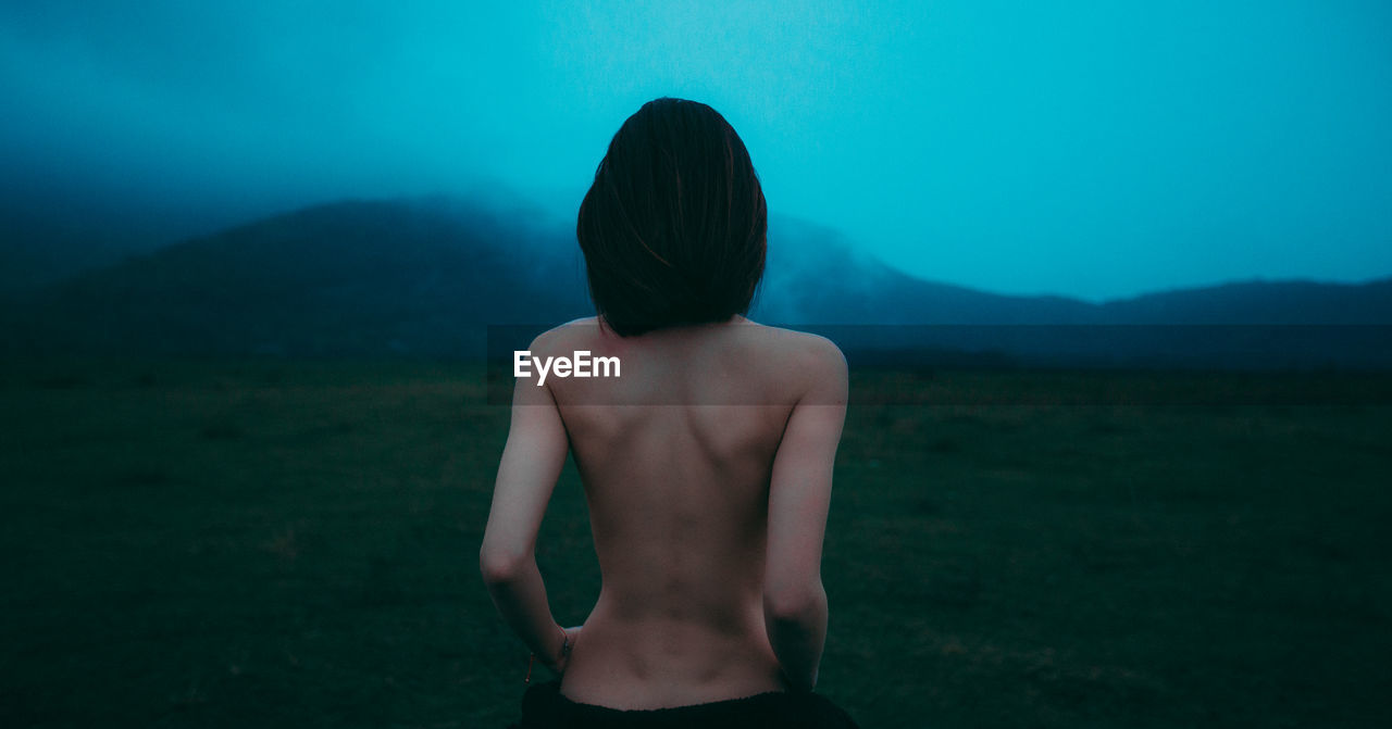 Rear view of shirtless woman on grass against mountain and sky during foggy weather