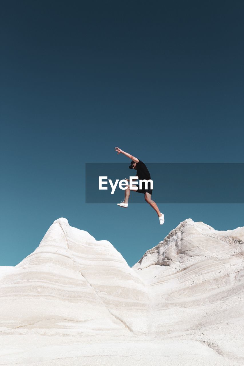 LOW ANGLE VIEW OF MAN JUMPING AGAINST MOUNTAIN