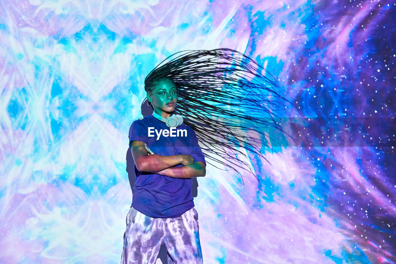 Side view of ethnic female millennial shaking long braided hair while his arms are crossed near wall with bright blue and lilac illumination