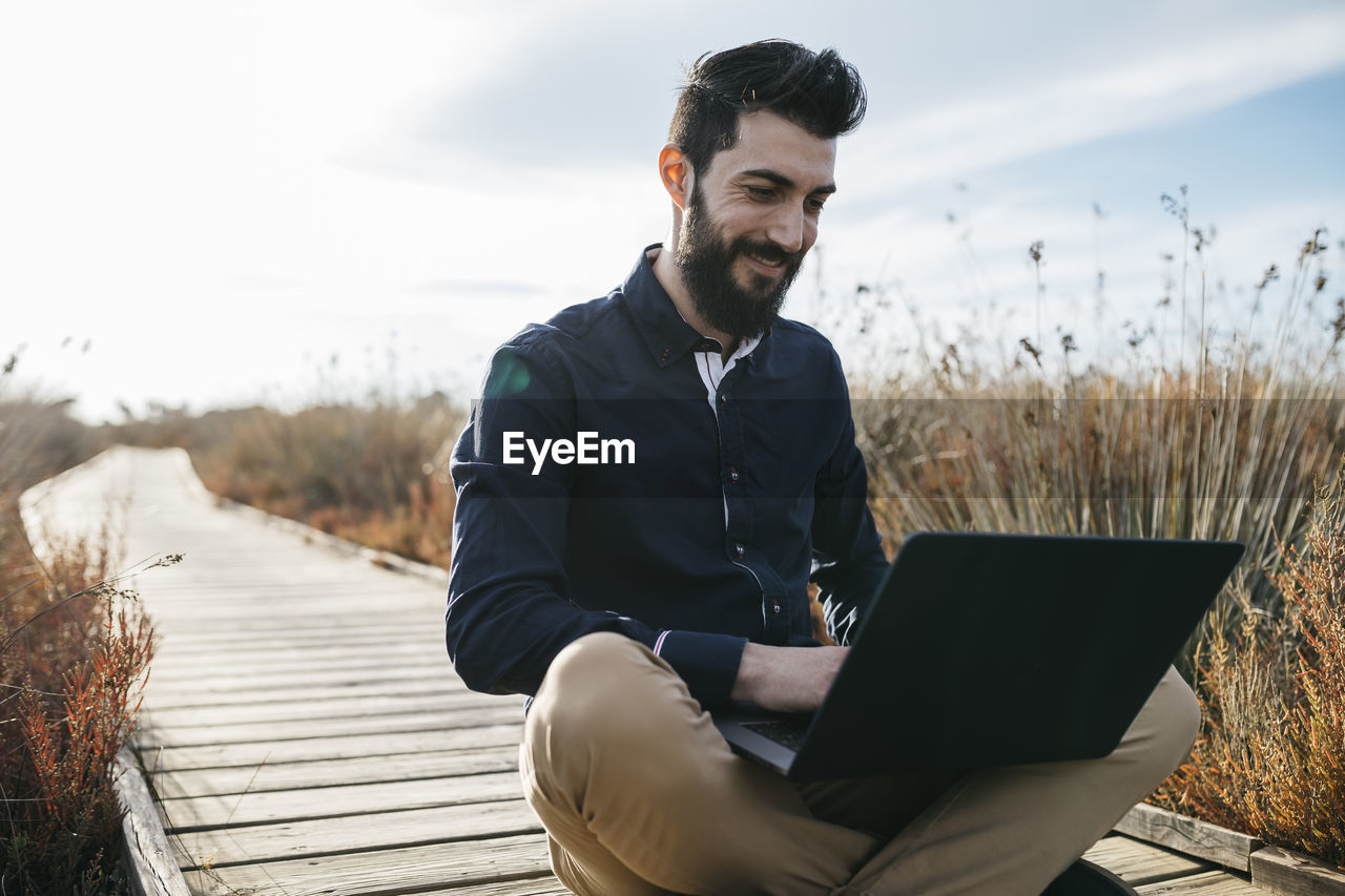 Man using laptop while sitting on boardwalk against sky