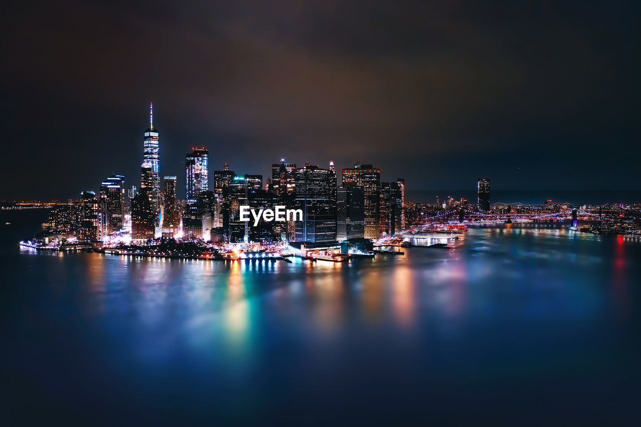Illuminated buildings reflecting on river against sky at night